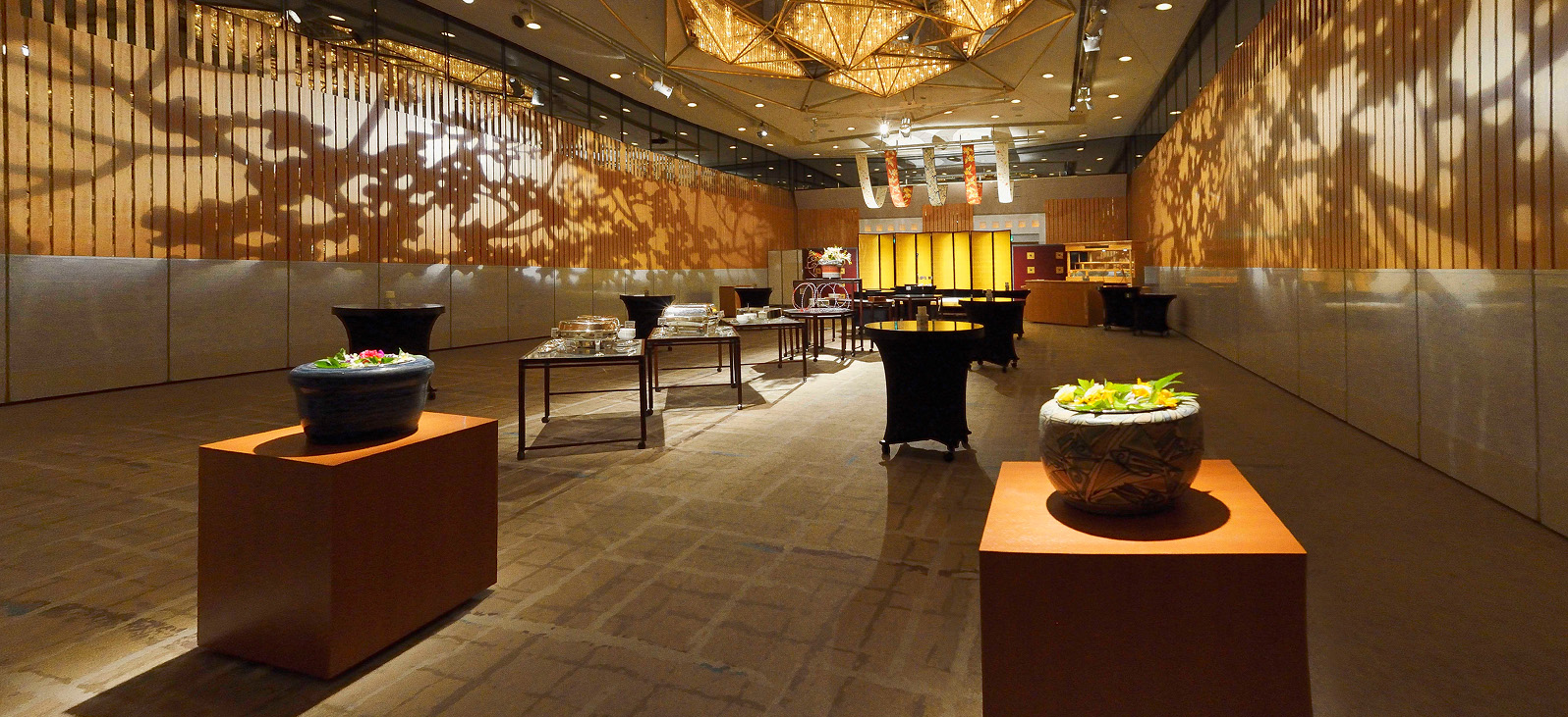 An overall view of the spacious event hall. On the front of the wood-grained wall, silhouettes of trees are projected onto most of the wall like shadow puppets. Several tables and chairs are arranged in the center of the room, and two decorated potted plants are placed in front of them.