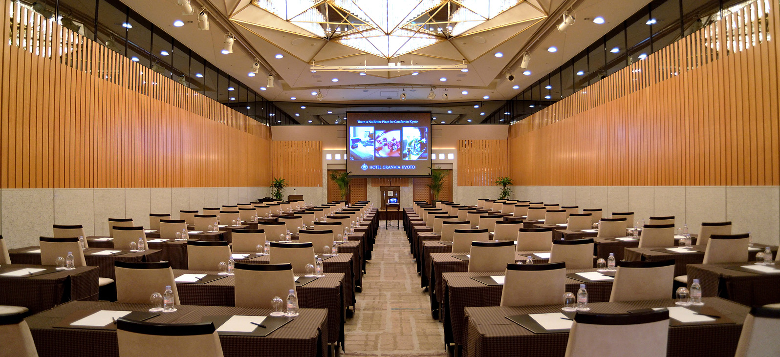 This is an overall view of the spacious Conference Rooms. In the center of the room, numerous conference tables and chairs are neatly arranged in a single row. A large screen is set up at the back of the room, and a platform resembling a podium can be seen below it. The walls are wood-grained, and the ceiling is made up of multiple panels with built-in lighting.