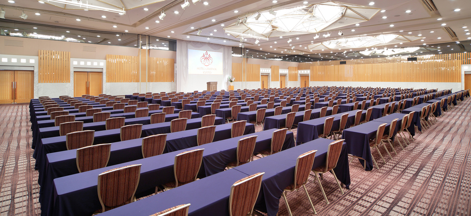 This is an overall view of the spacious event hall. From the front to the back, a large number of long tables and chairs are neatly arranged. The tables are covered with navy blue tablecloths, and the chairs feature wooden backs and seats. At the back of the room, a white screen is set up, displaying red patterns and text. The walls of the room have a wood-grain finish, and multiple lights are positioned above.