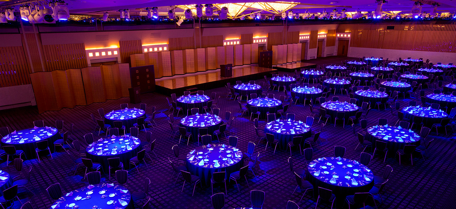This is an overall view of the spacious event hall, adorned with dazzling lighting. The interior is primarily illuminated with purple and blue lights, creating a fantastical atmosphere. Numerous round tables are arranged from the front to the back, each illuminated with blue light, creating white patterns on their surfaces.