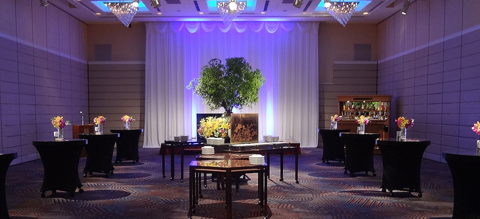 The event venue features a white curtain illuminated by blue lights, and a large bonsai plant is displayed in the center. The venue features round tables covered in black cloth and several wooden tables, decorated with colorful floral arrangements. In the back right, you can see a bar counter lined with alcoholic beverages.
