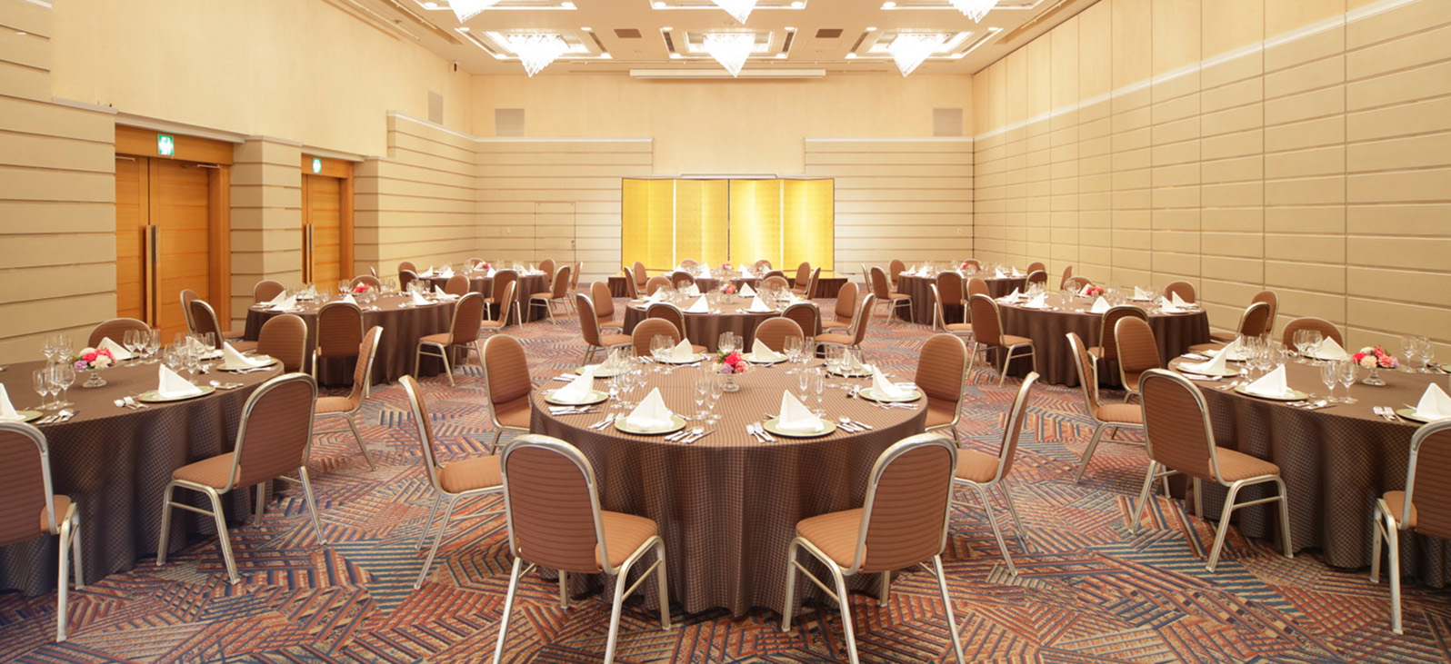The spacious banquet hall was filled with several round tables and chairs neatly arranged. Each table was set with white tableware and glasses, pink napkins, and adorned with a small floral arrangement. The walls of the hall were beige, and numerous chandeliers and downlights were installed on the ceiling.