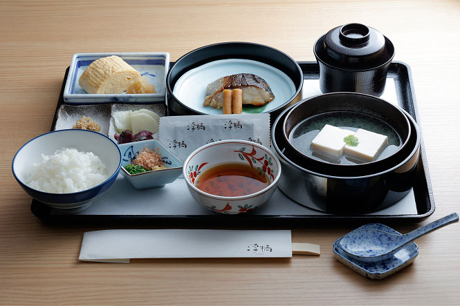 A Japanese breakfast set. Grilled fish, tofu, omelet, small side dishes, rice, miso soup, and other Japanese dishes are neatly arranged.