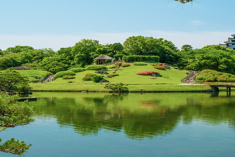 穏やかな池の水面に、緑豊かな築山や木々、小さな東屋が美しく映り込んでいる、晴天の日本庭園の風景。