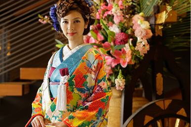 色鮮やかな振袖を身にまとった女性が、華やかな花々を背景にして微笑んでいます。