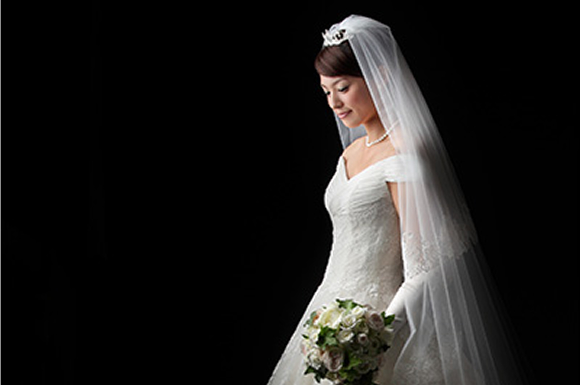 白いアイアンゲートの向こうで、優しく手をつなぎ見つめ合う新郎新婦の後ろ姿。レンガ調の壁に囲まれた空間で、正面の大きな格子窓から差し込む眩い光に包まれた、ドラマチックで美しいウェディングフォトの一枚。