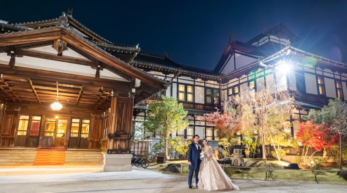 灯りが灯った本館を背景に寄り添う洋装姿の新郎新婦