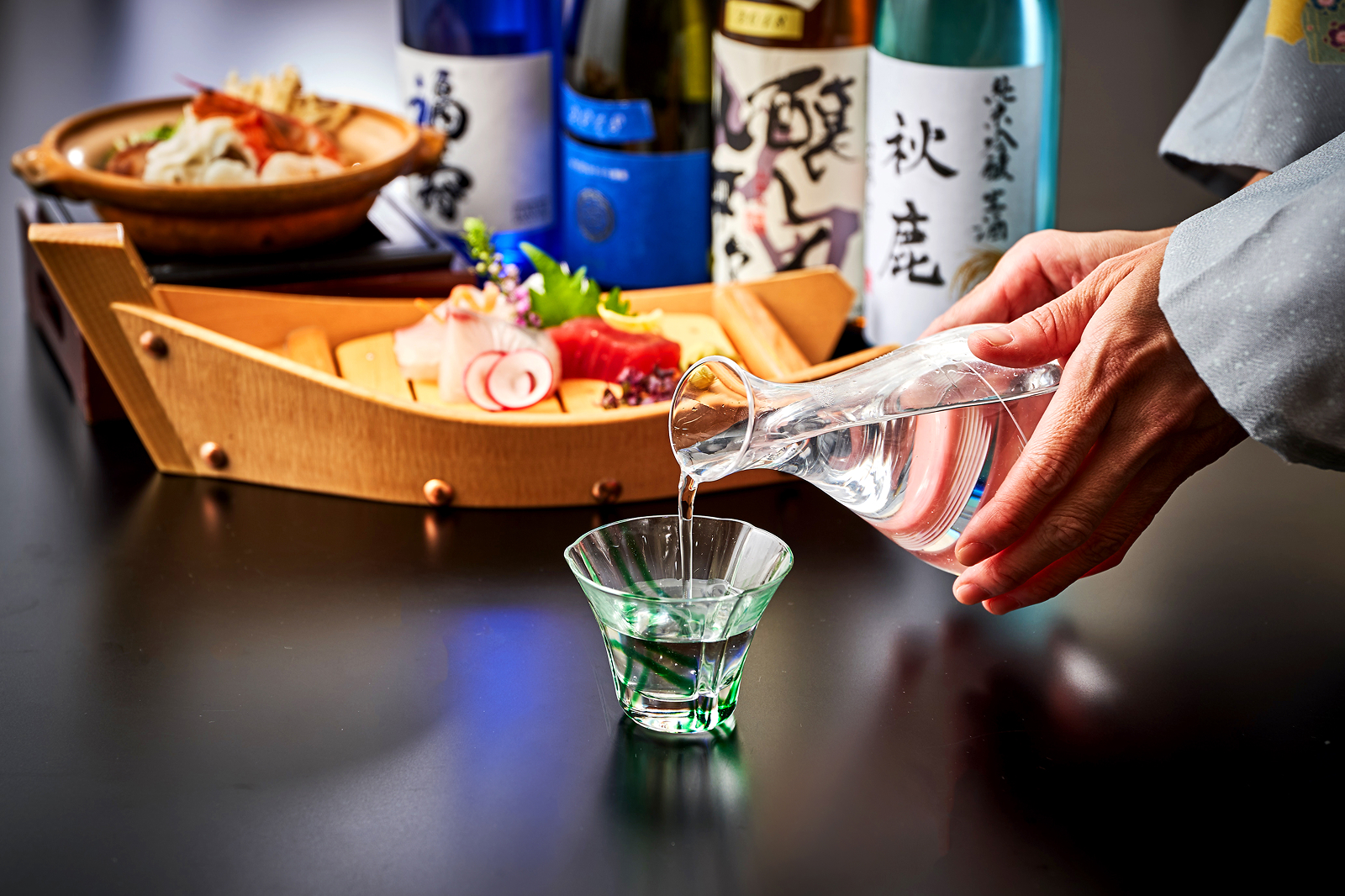透明な徳利からガラスの猪口へ日本酒が注がれ、背景には舟盛りの刺身と銘酒の瓶が並ぶ。