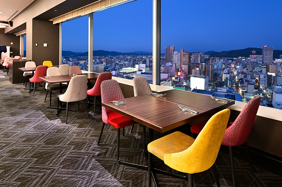 2階層分の高さがある巨大な全面窓から夜景を一望できる、圧倒的な開放感のダイニング＆バー。手前には緩やかな曲線を描く艶やかな赤茶色の大型カウンターと白い椅子が並び、ガラス窓に映り込む青いイルミネーションが都会的でお洒落な雰囲気を醸し出す店内の様子。